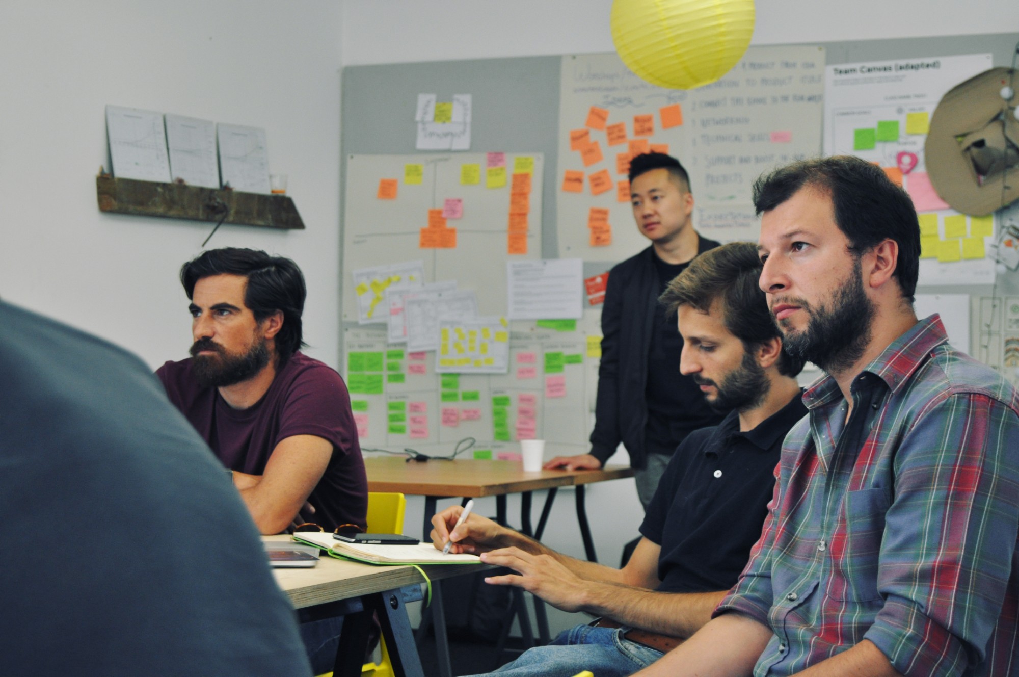 Mentors and students listen to a project presentation, with Verne Ho standing in front of boards covered in sticky notes