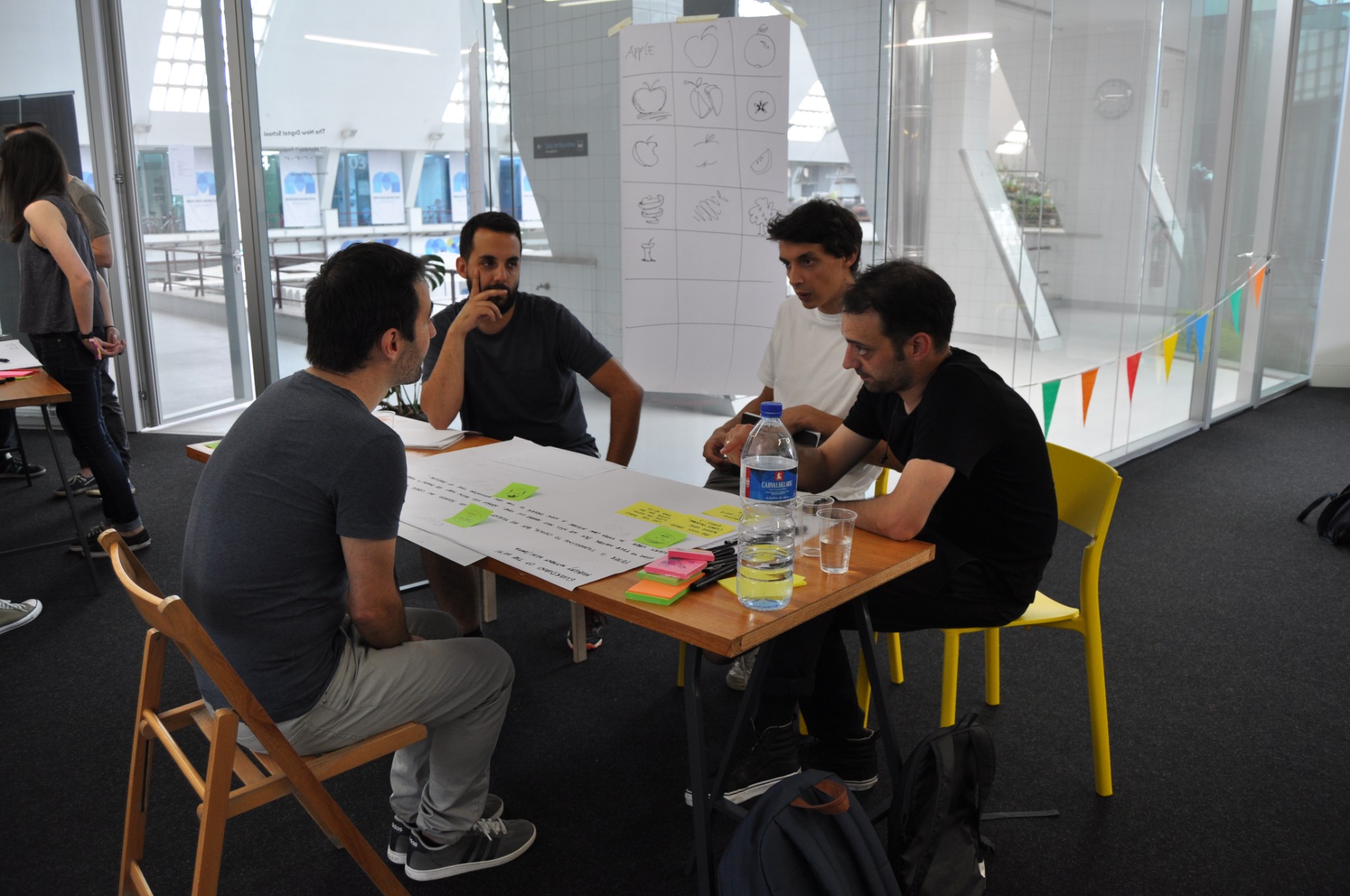 Four workshop participants discuss ideas over sticky notes, with a sheet of apple sketches taped to the glass behind them