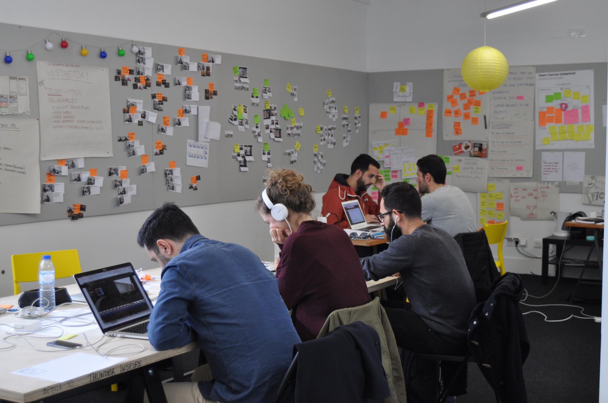 Students work on laptops at long tables beneath a wall of research photos, sticky notes and project briefs